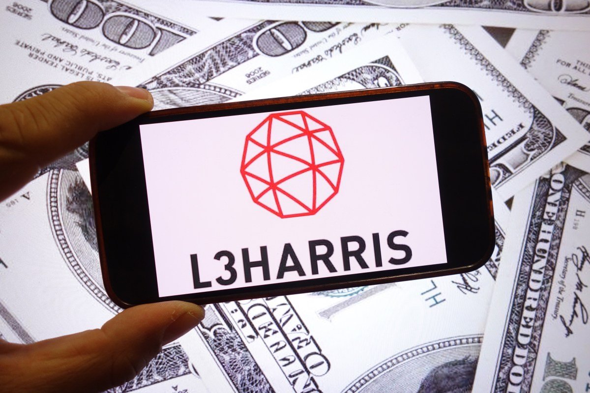 The L3Harris Technologies company logo is seen displayed on a smartphone screen with dollars in the background.